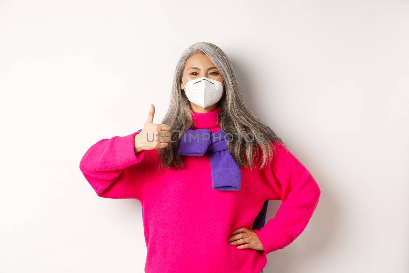 Covid, pandemic and social distancing concept. Cheerful asian senior woman in respirator showing thumbs-up, wearing face mask from coronavirus, white background by Benzoix