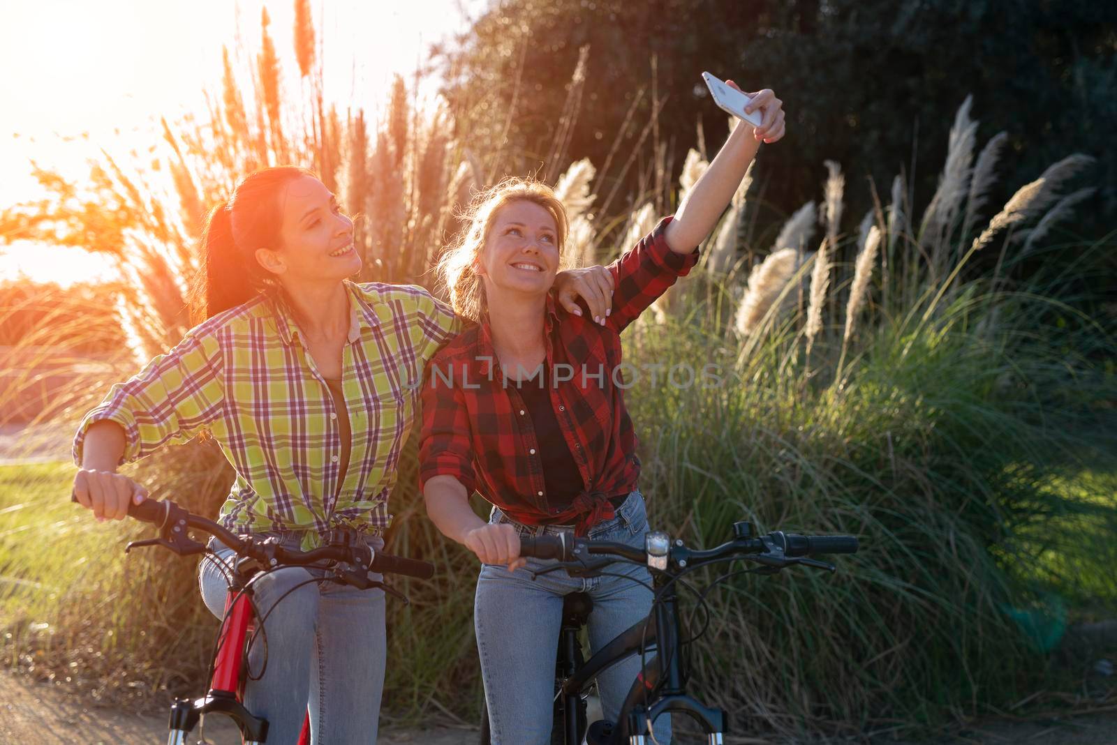 Two pretty young caucasian girls making selfie photo with smartphone. Best friends enjoying a day on bikes. Sunny summer evening.