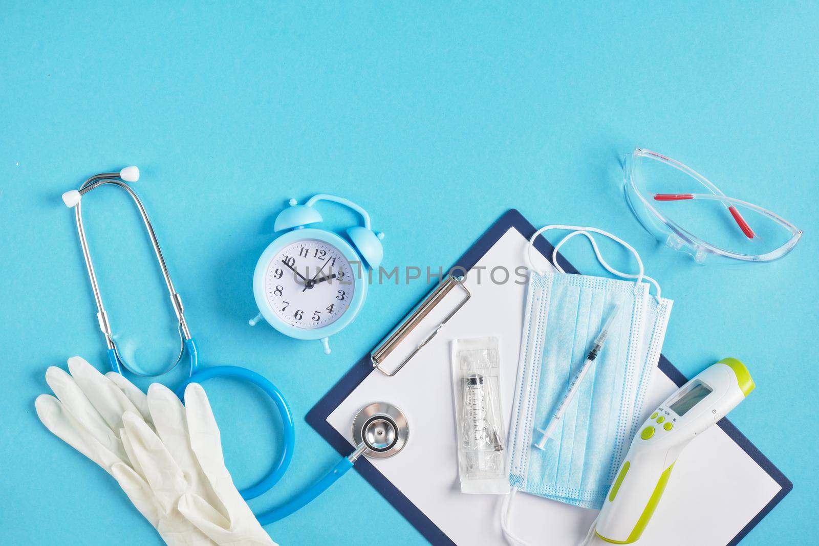 Alarm clock and medical equipment on a blue background place copy top view stethoscope non-contact thermometer face mask syringe safety glasses and gloves