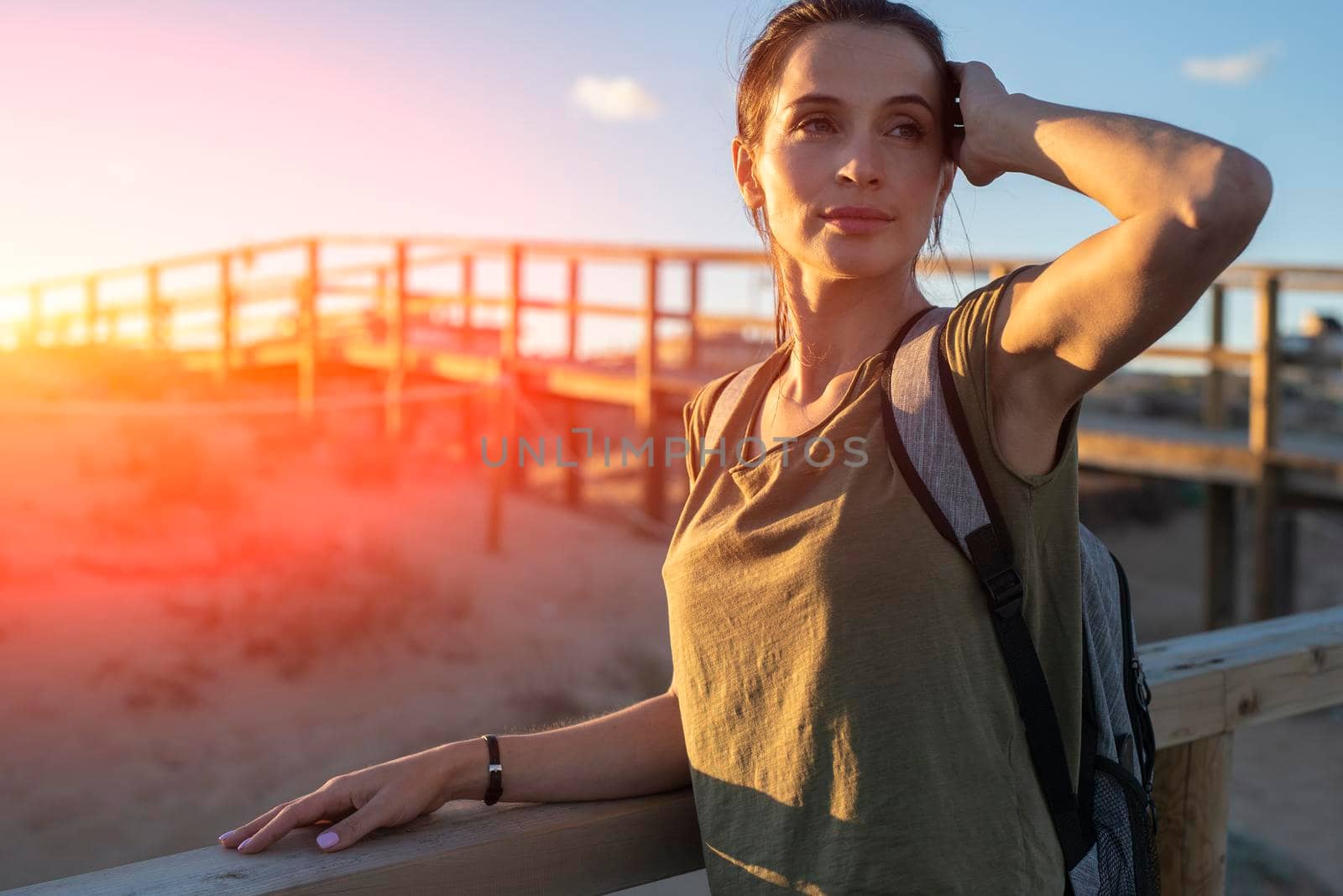 Fashionable slender young brunette girl with backpack posing on a background wooden walkways and sand beach at sunset.