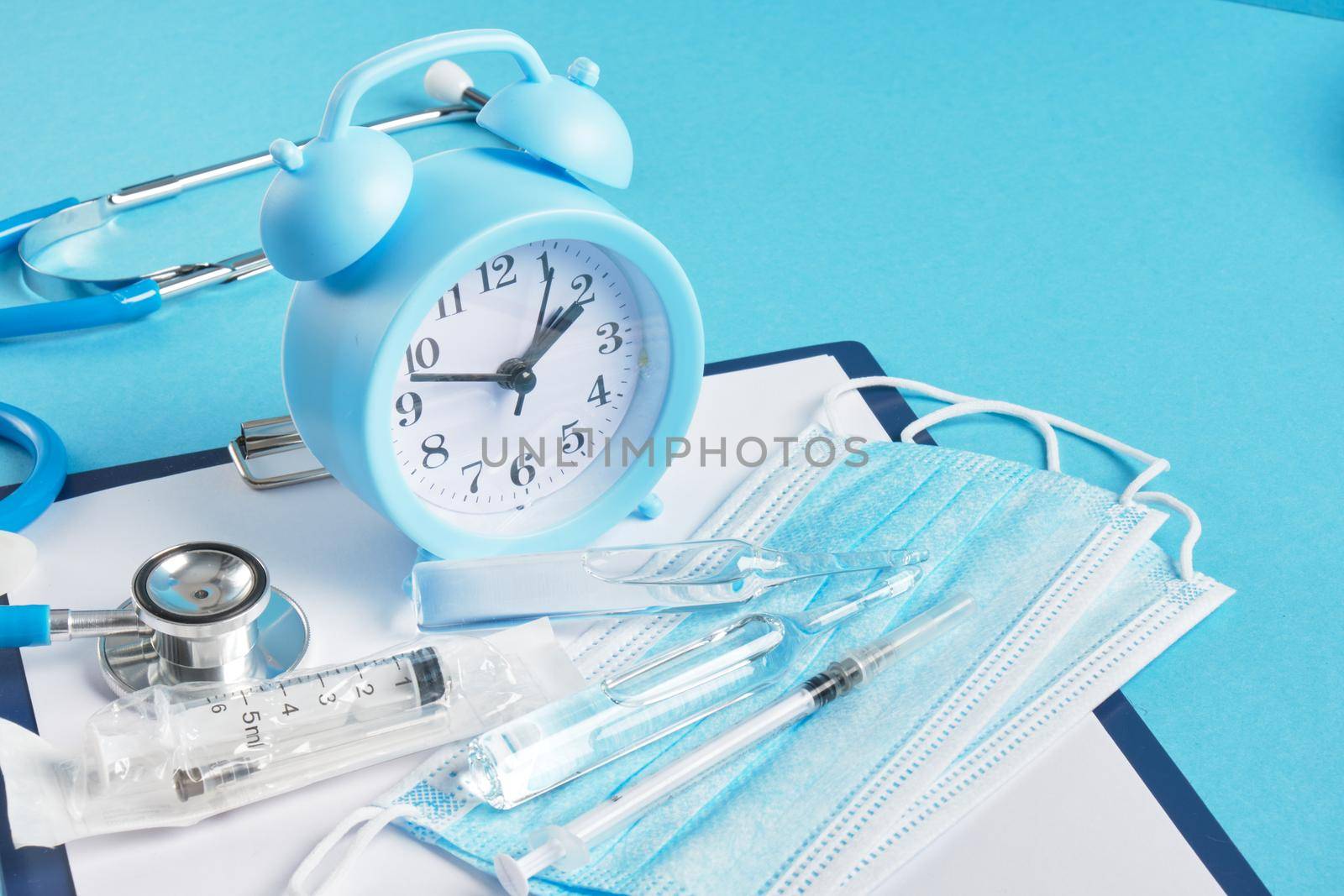 Alarm clock and medical equipment on blue background copy space top view stethoscope non-contact thermometer ampoule face mask syringe safety glasses
