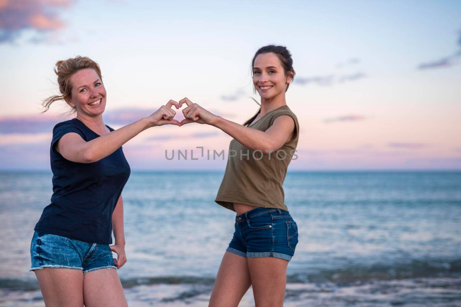 leisure and friendship concept - two happy smiling teenage girls or best friends at seaside making hand heart gesture. selective focus.