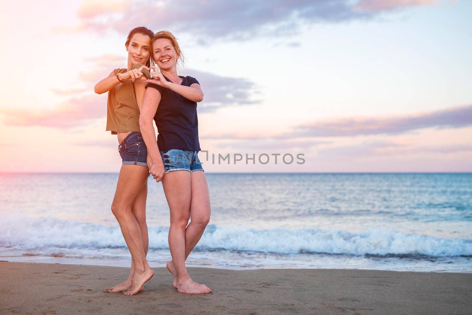 leisure and friendship concept - two happy smiling teenage girls or best friends at seaside making hand heart gesture at sunset.