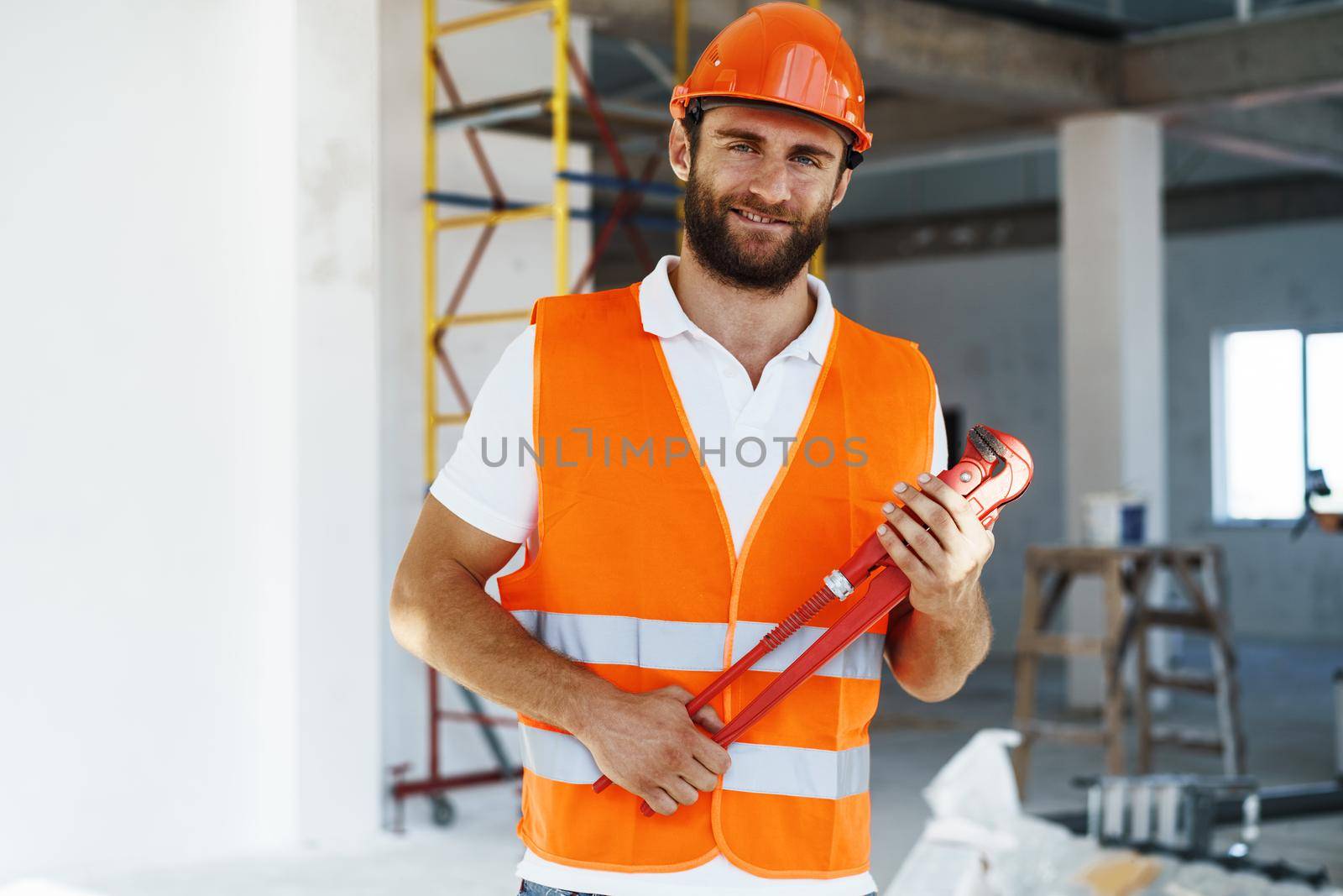 Young male plumber in workwear holding pipe wrench on a construction site indoors