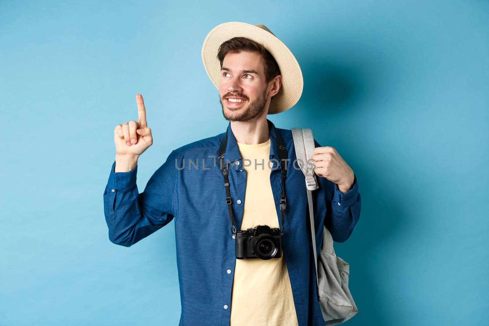 Handsome positive guy on vacation, wearing summer hat, looking and pointing up at logo, smiling pleased, standing with camera and backpack on blue background by Benzoix