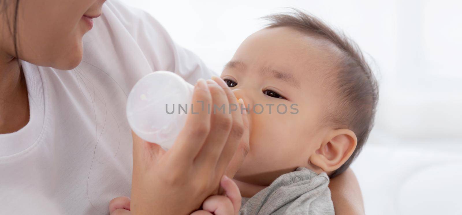 Young asian mother embracing and feeding little baby girl with bottle of milk at home, newborn innocence drinking with mom satisfied, relationship and bonding of mum and child, family concept.