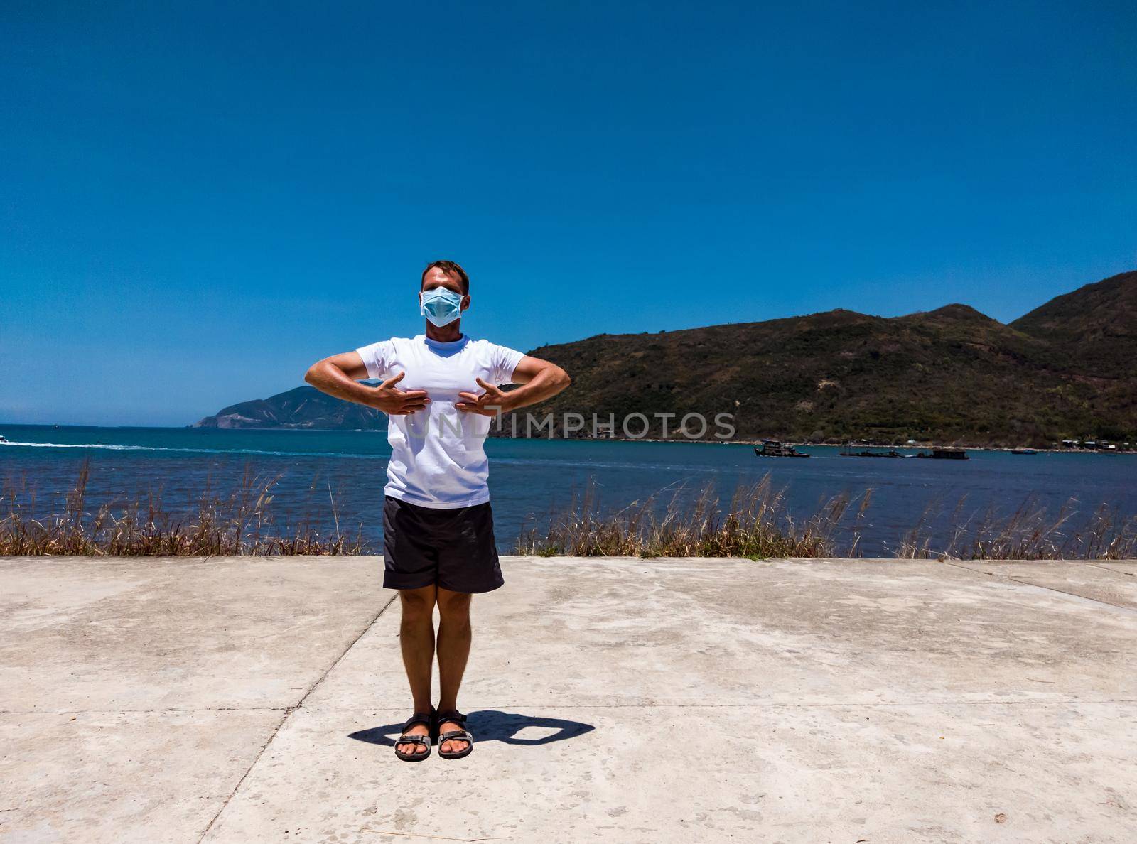 Coronavirus COVID 19. Young beautiful man doing sport performing workouts exercises in the park near sea during quarantine. Active life in surgical sterilizing face mask protection.