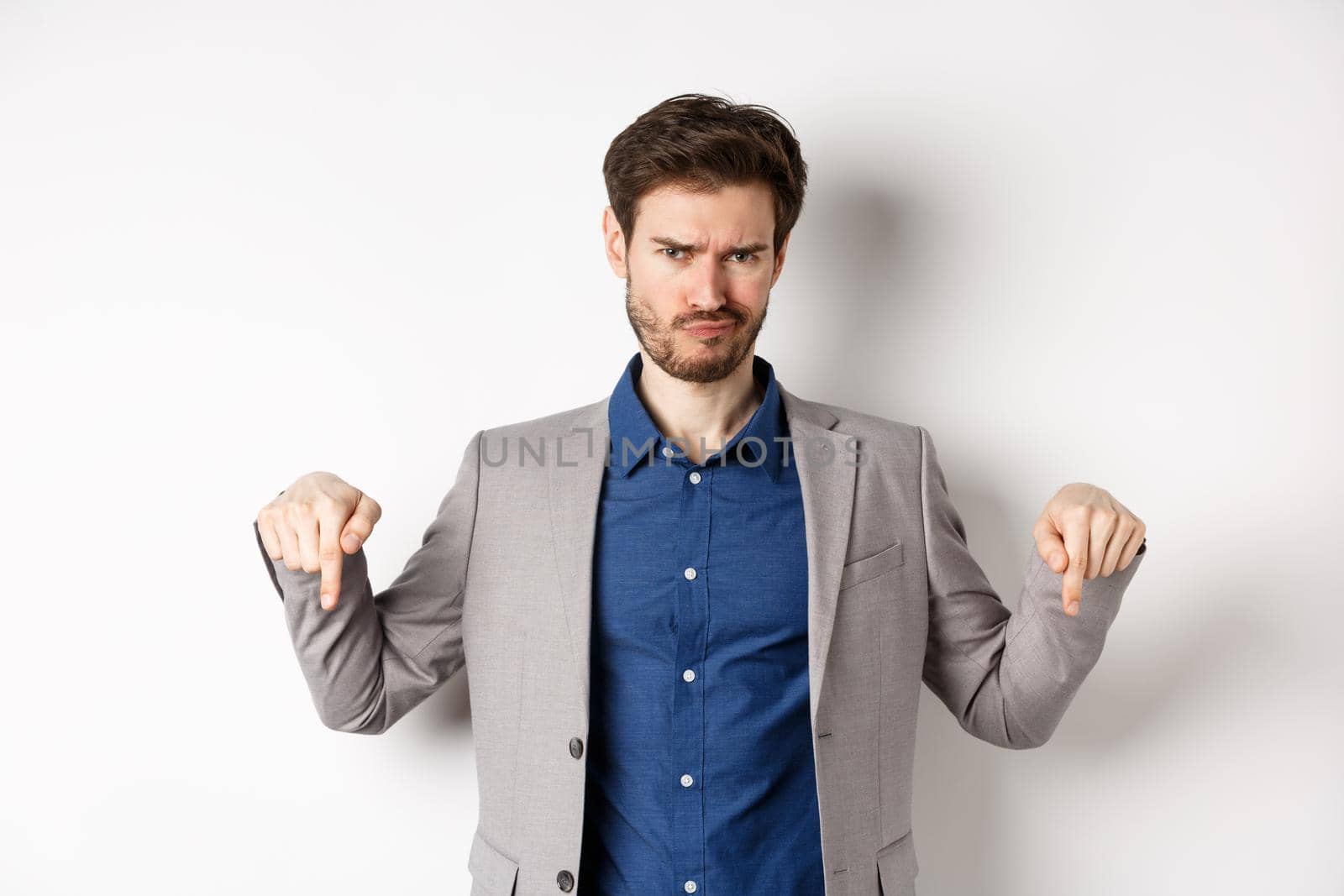 Disappointed bearded male entrepreneur frowning and pointing down with negative emotion, express dislike or bad opinion, standing on white background.