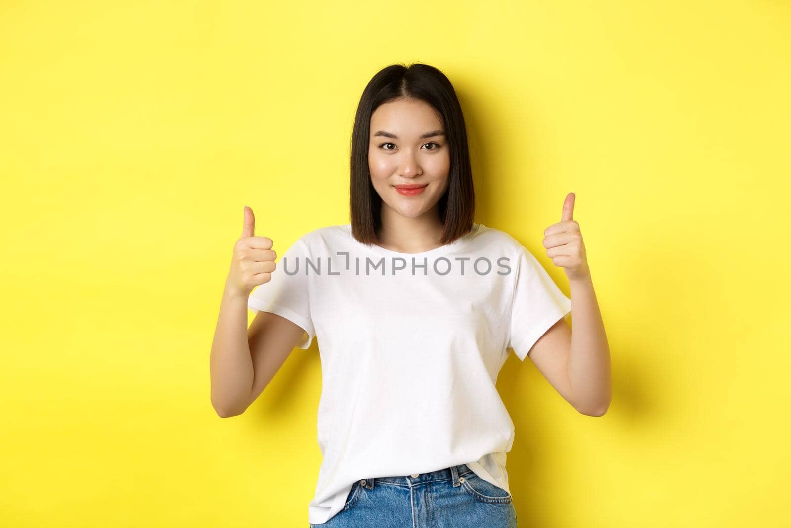 Pretty young asian woman in white t-shirt, showing thumbs up and smiling, praise good offer, recommend product, standing satisfied over yellow background by Benzoix