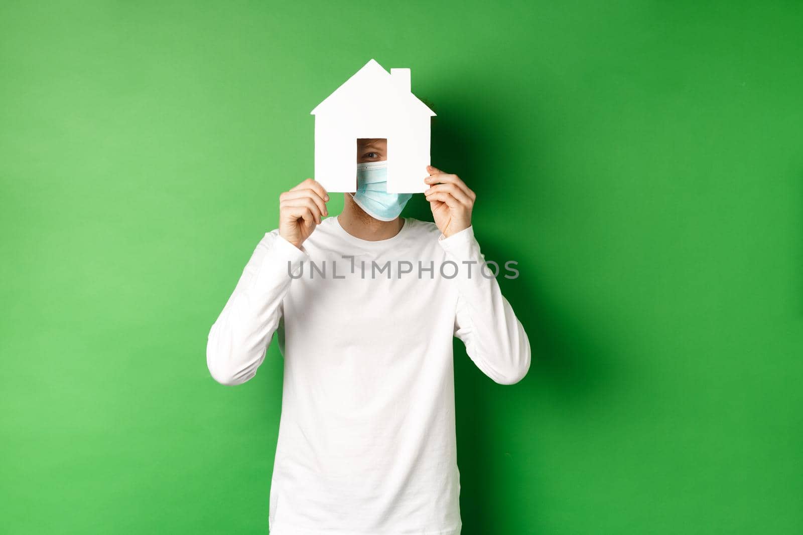 Real estate and covid-19 pandemic concept. Funny young man in face mask and white long-sleeve hiding face behind paper house cutout, peeking at camera, green background by Benzoix