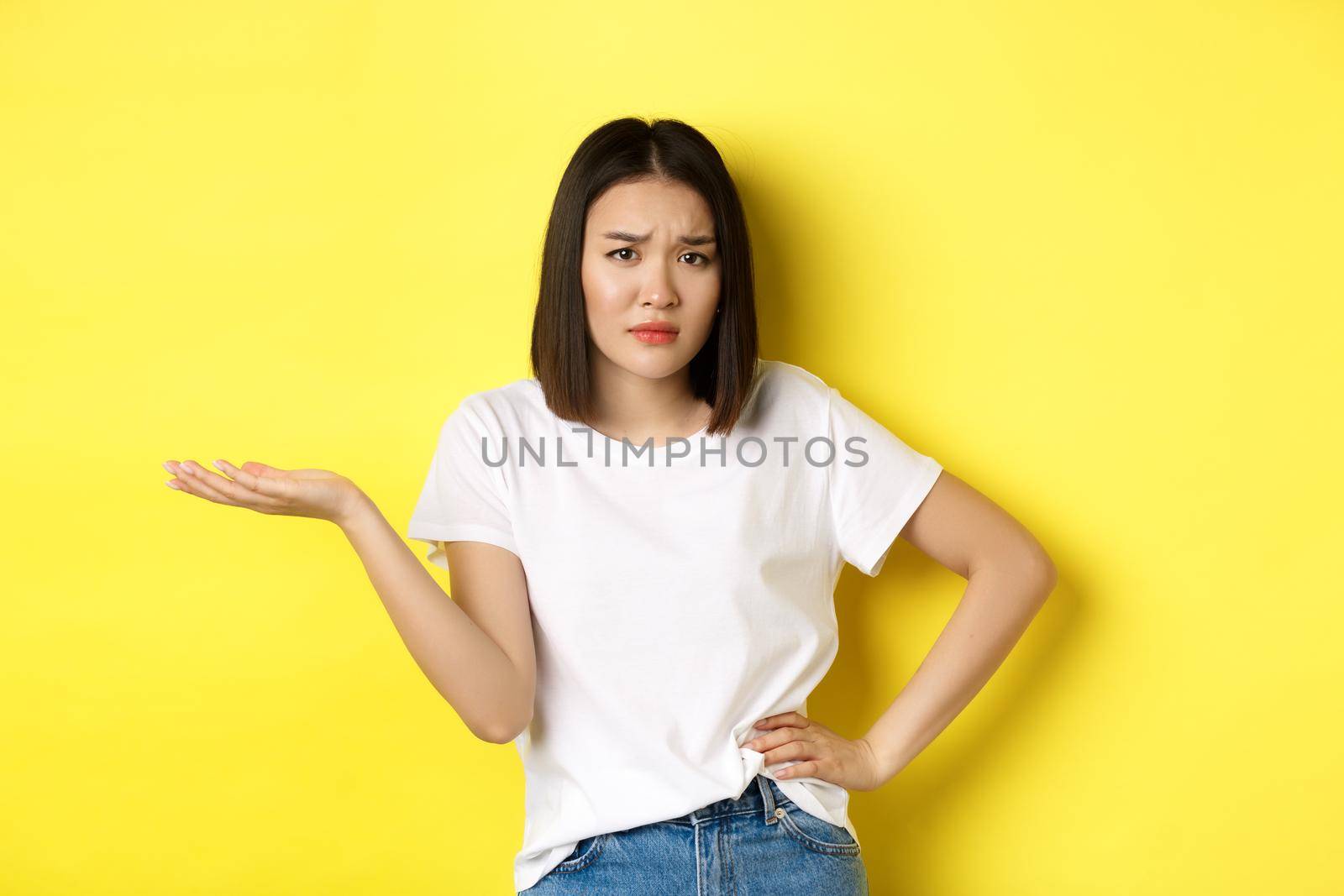 Disappointed asian woman asking so what, raise hand up and staring skeptical at camera, standing over yellow background by Benzoix
