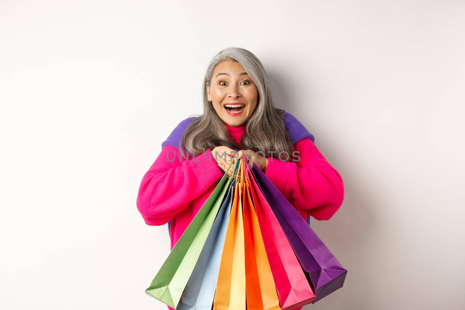 Fashionable senior asian woman shopaholic, hugging shopping bags and smiling joyful, buying with discounts, standing over white background by Benzoix
