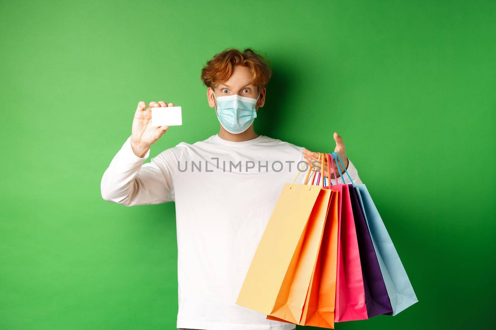 Handsome young man in medical mask, showing plastic credit card and shopping bags with items purchased with discount, green background by Benzoix