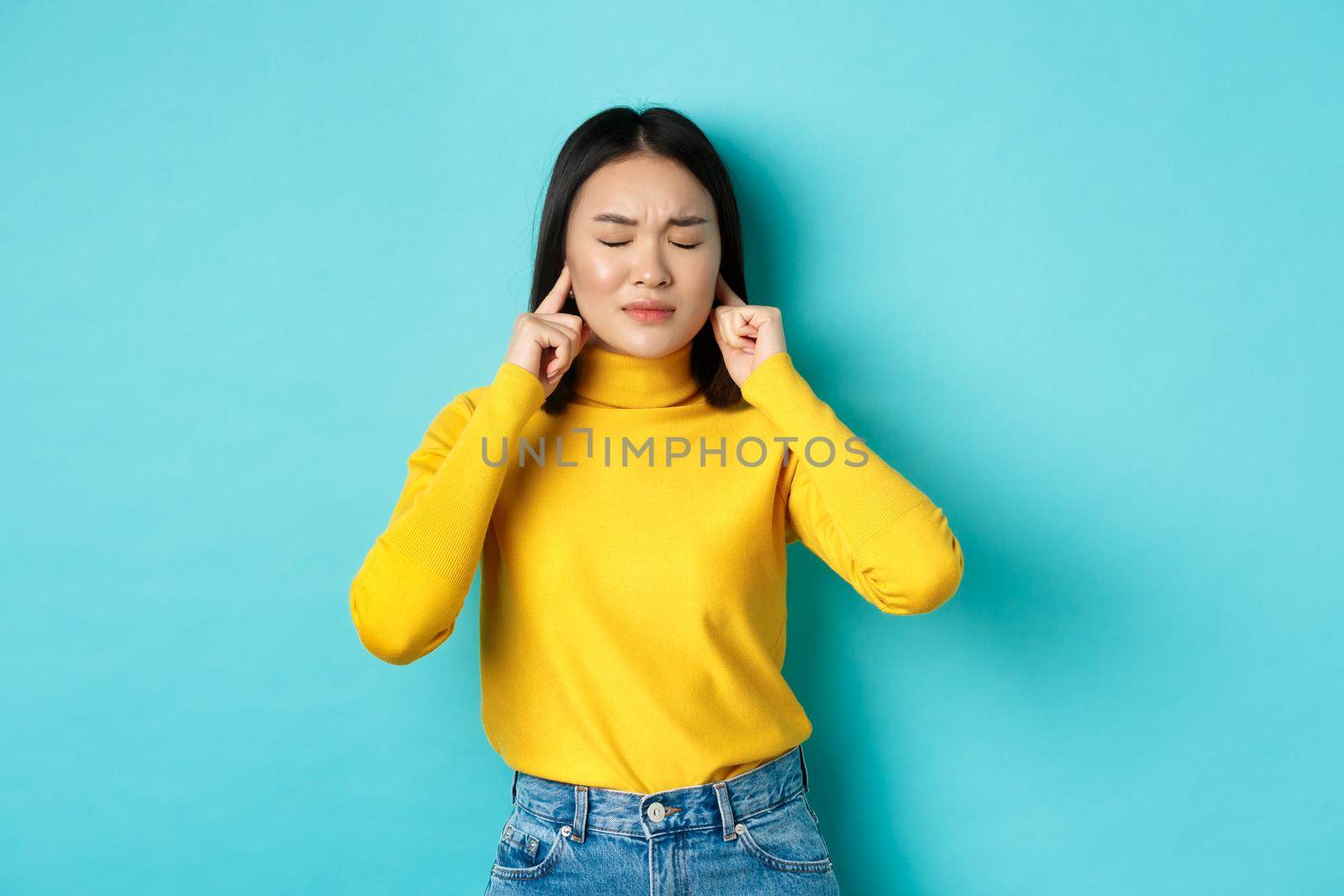 Tired and disappointed young asian woman unwilling to listen, shut ears with fingers and close eyes, standing in denial over blue background.
