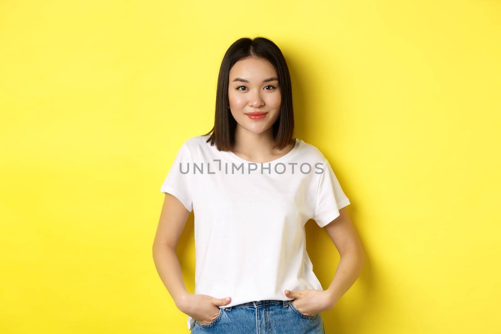 Confident and stylish asian woman holding hands on jeans and smiling, standing over yellow background.
