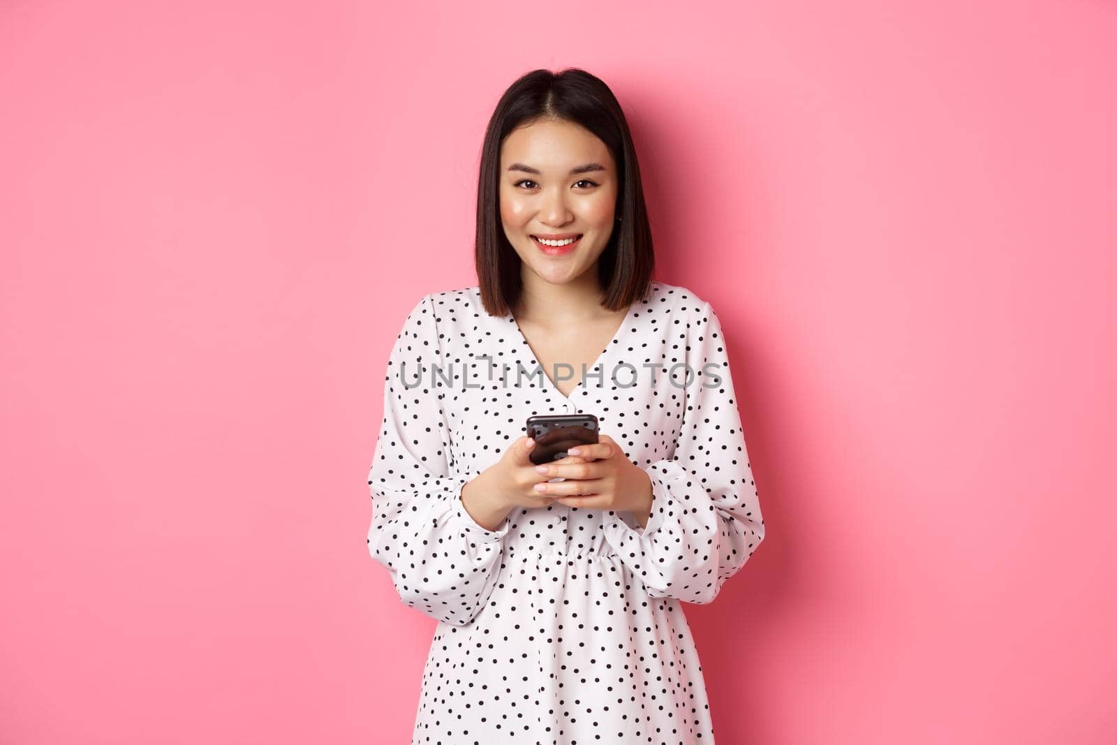 Beautiful asian lady reading message and smiling, using mobile phone, standing in cute dress against pink background.