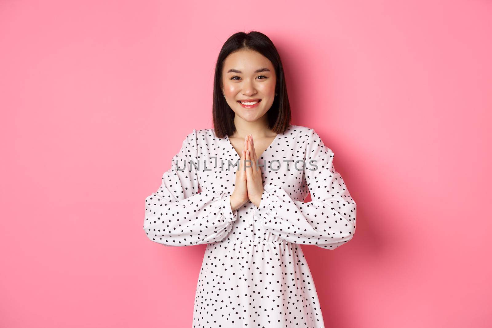 Beautiful asian woman thanking you, holding hands together in appreciation gesture, smiling happy at camera, standing grateful over pink background by Benzoix