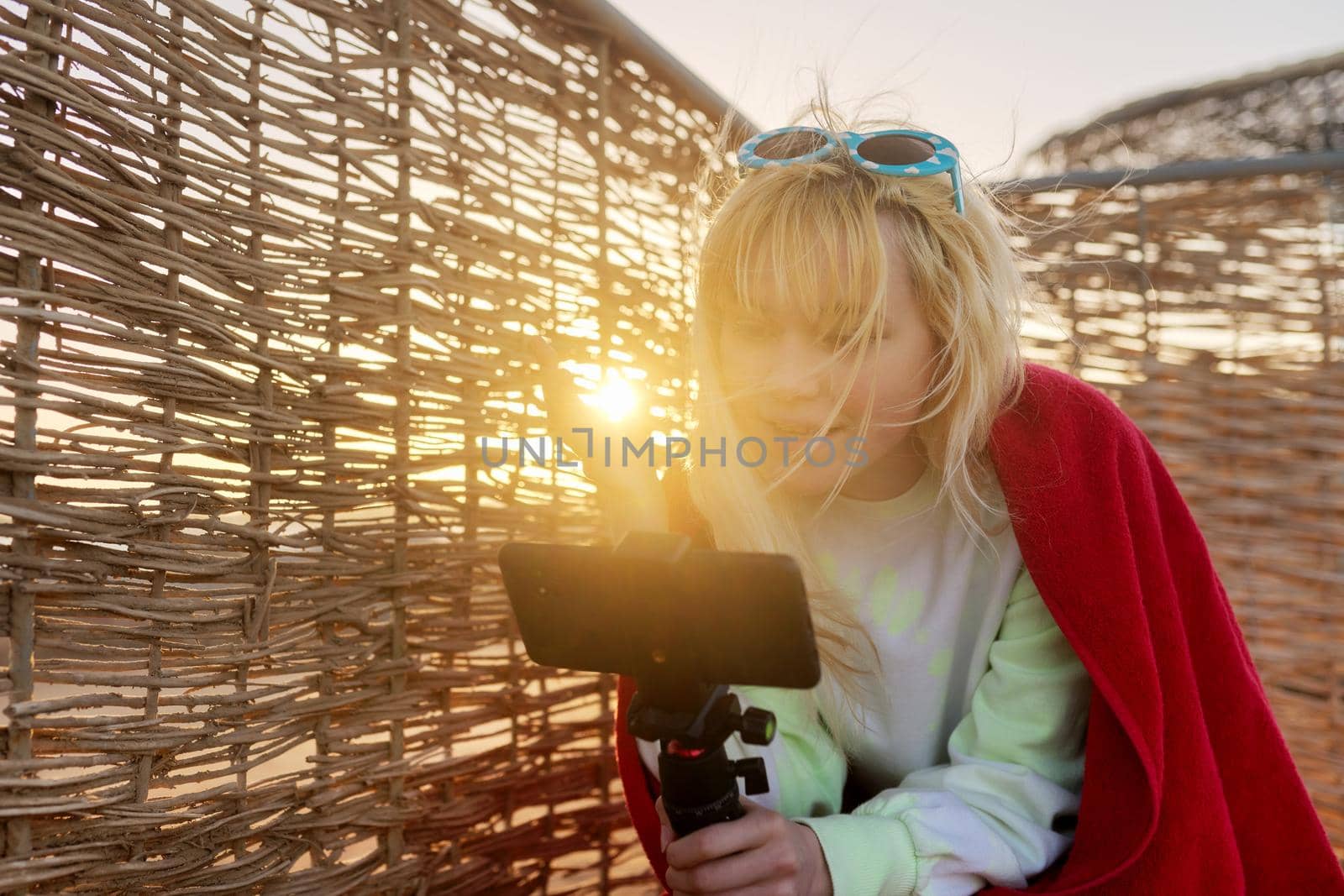 Outdoor sunset portrait of female teenager recording video on smartphone, rattan, sky background. Video call, stream, blog, vlog, telecommunications technologies, youth concept, copy space