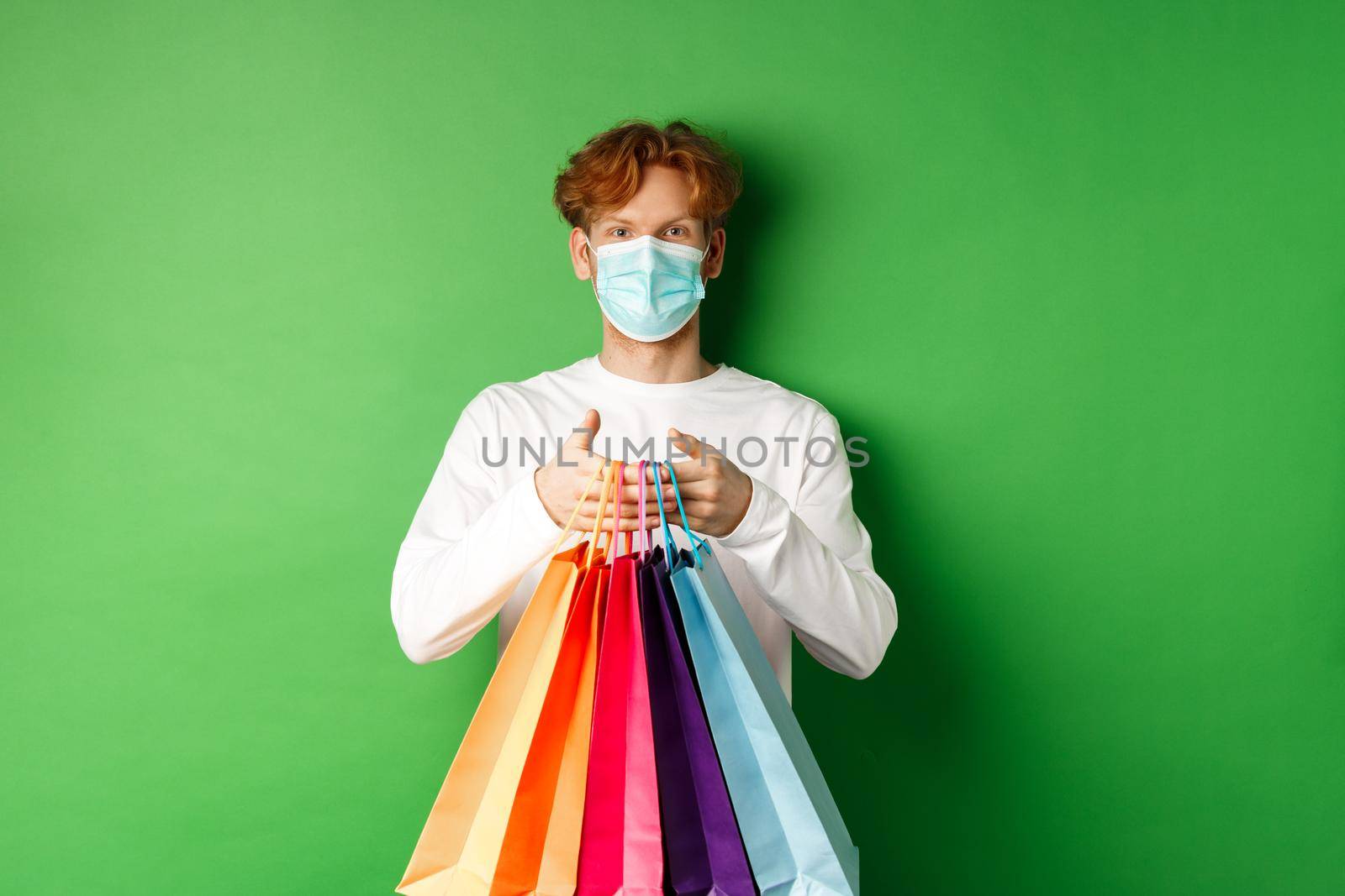 Pandemic and lifestyle concept. Cheerful redhead man going shopping in store, wearing medical mask and holding bags, standing over green background.