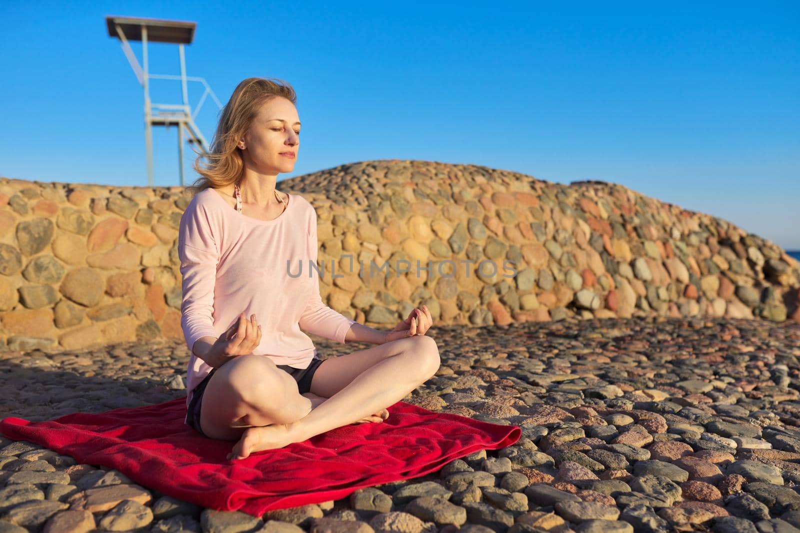 Beautiful mature woman blonde sitting in lotus position and meditating on the seashore at sunset. Relaxation, relaxation, enjoying nature by the sea