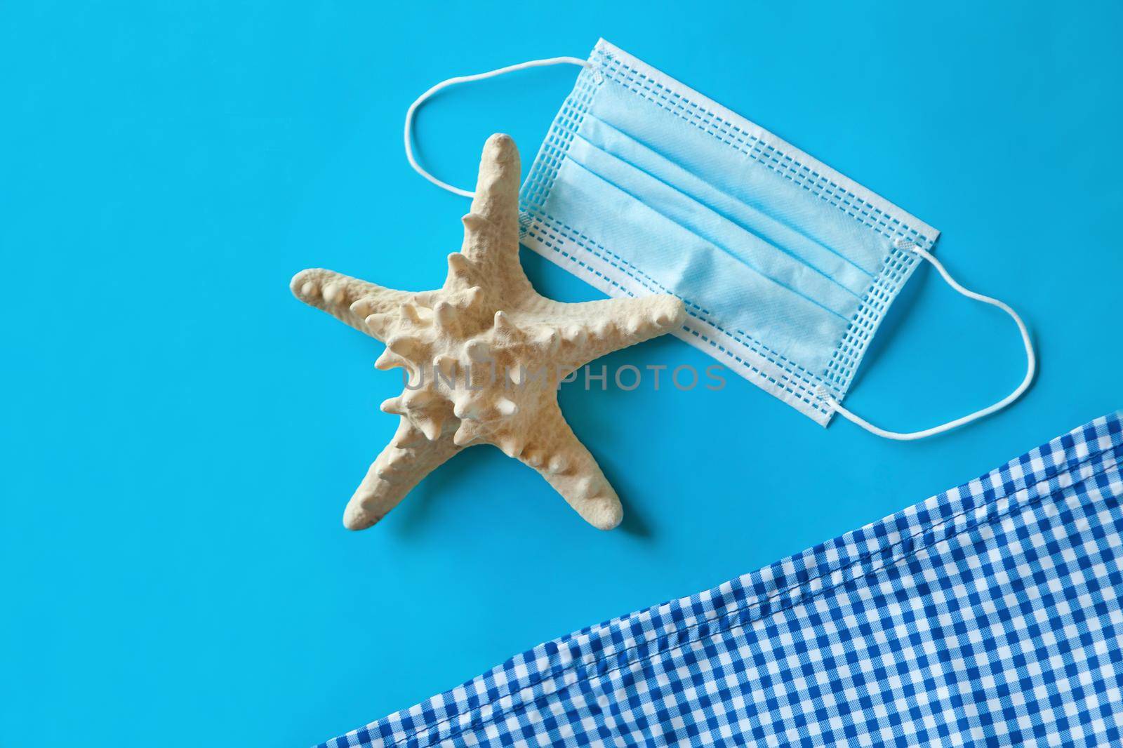 Medical face mask near beach towel and starfish on a blue background, top view, beach items and summer vacations concept