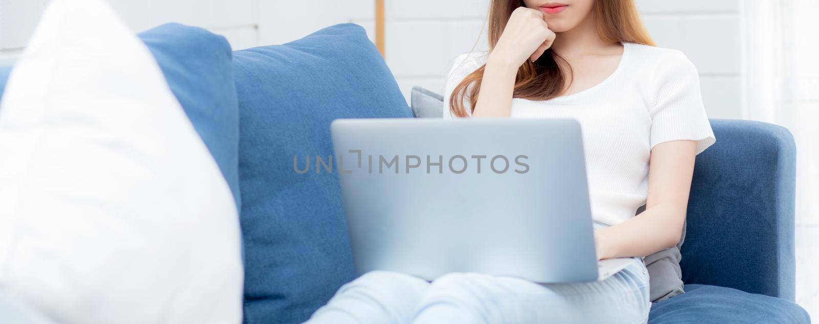 Young asian business woman smile and work from home with laptop computer online to internet on sofa in living room, freelance girl using notebook on couch with comfort, new normal, lifestyle concept.