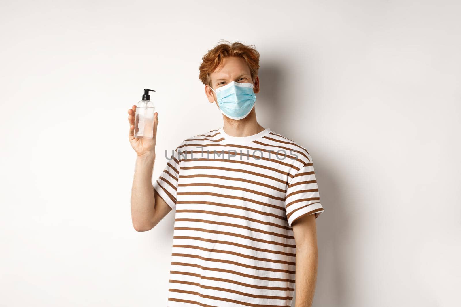 Covid-19, health and lifestyle concept. Smiling male model with red hair, wearing face mask, showing hand sanitizer, recommend to use antiseptic, white background.