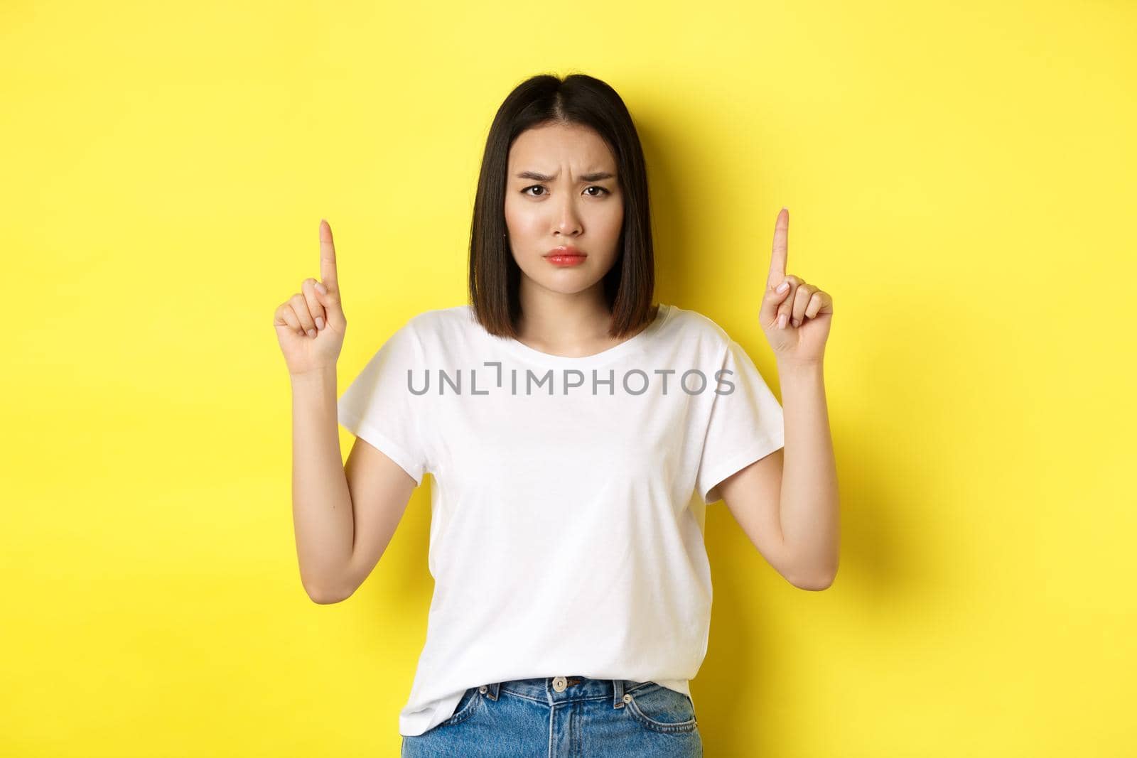 Beauty and fashion concept. Beautiful asian woman in white t-shirt pointing fingers up, standing over yellow background by Benzoix
