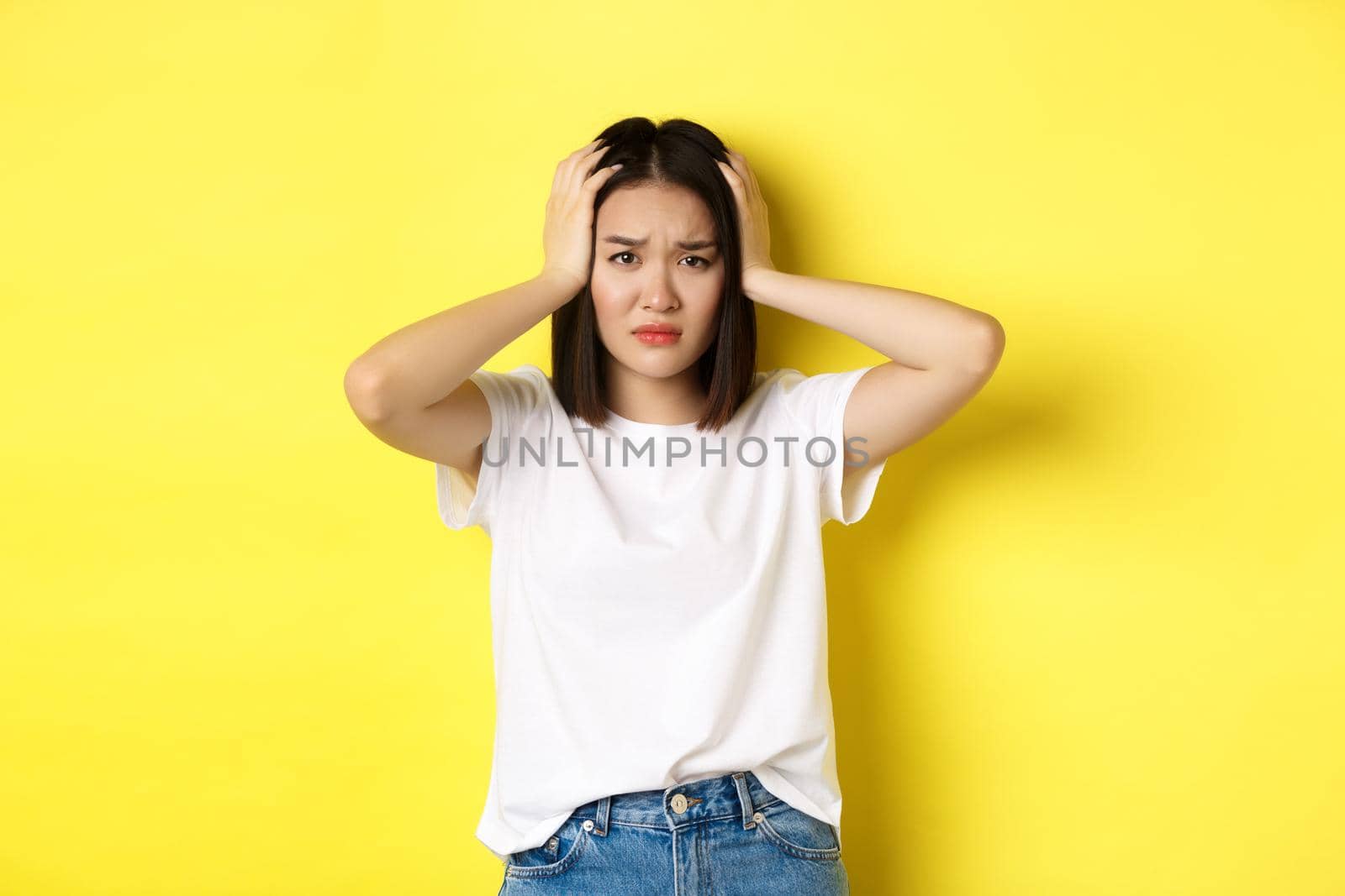 Asian woman holding hands on head and looking sad, having a problem, standing anxious against yellow background.
