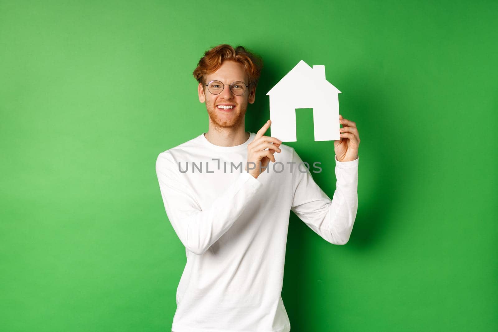 Real estate. Handsome young man with red hair, wearing glasses, showing paper house cutout and smiling, standing against green background by Benzoix