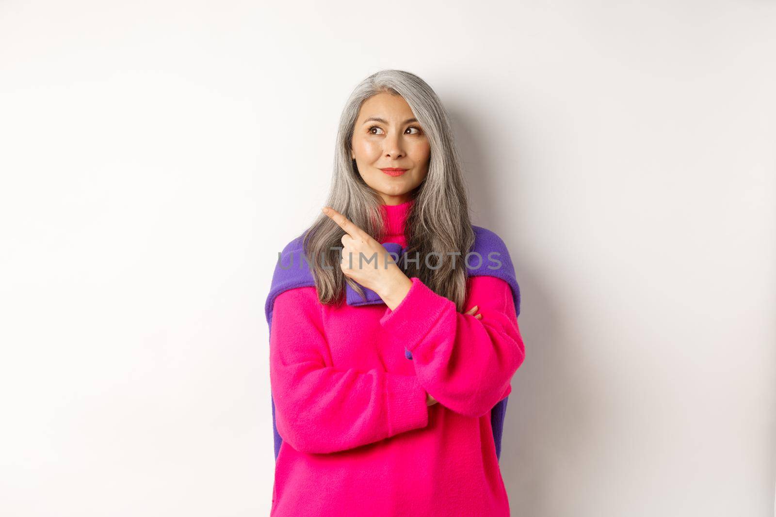 Stylish modern asian elderly woman pointing at upper left corner, looking at logo with pleased smile, standing in pink sweater over white background by Benzoix