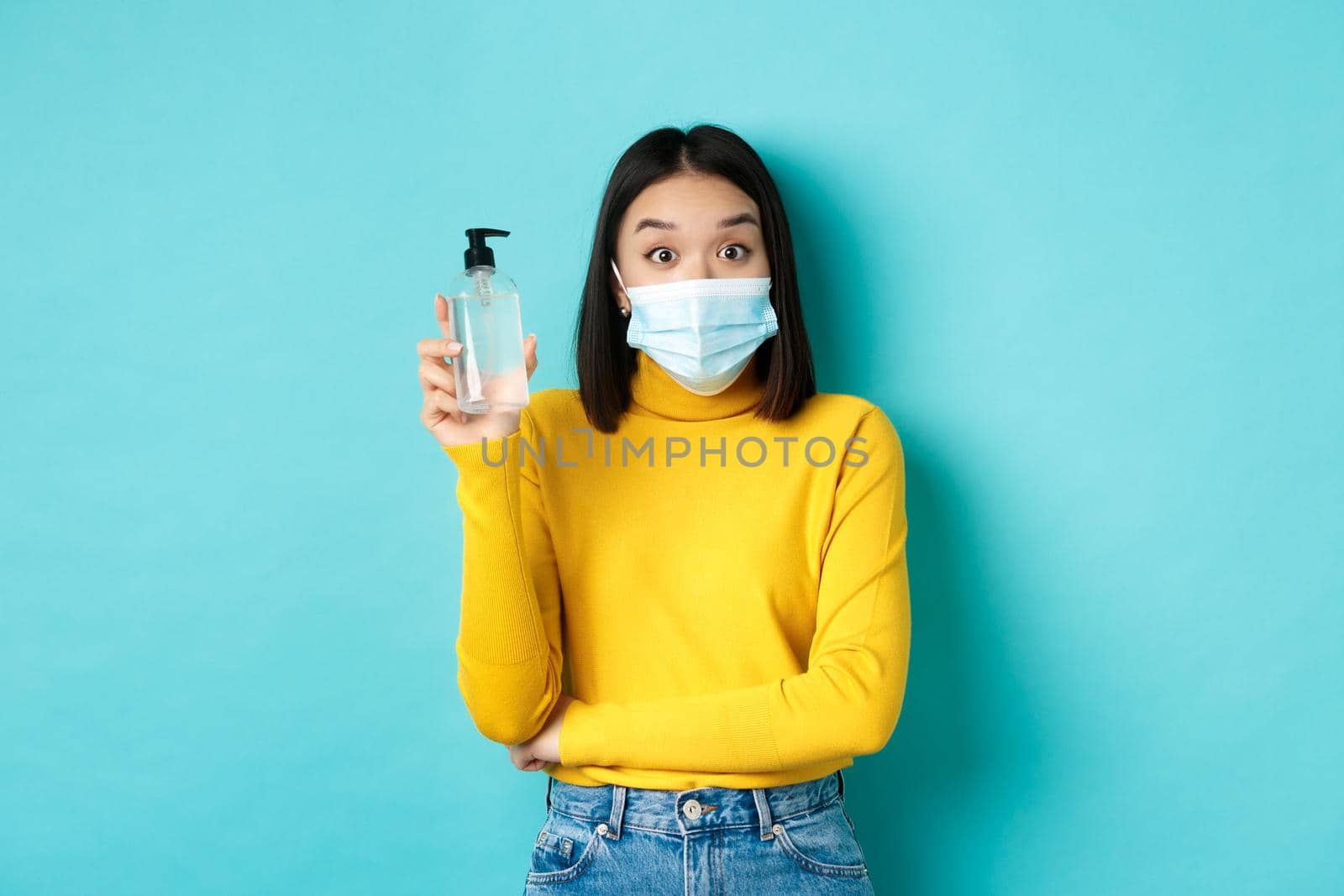 Covid-19, social distancing and pandemic concept. Cheerful asian woman showing hand sanitizer, recommend use antiseptic from coronavirus, wearing medical mask by Benzoix