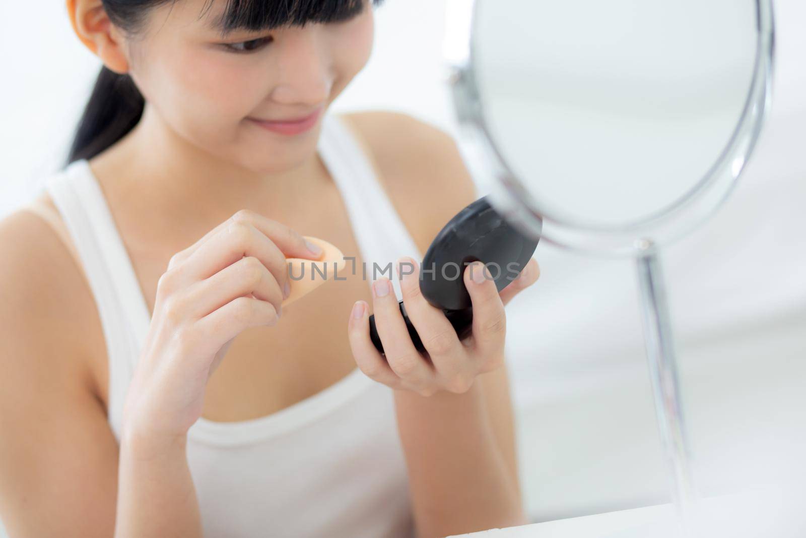 Beautiful young asian woman applying powder puff on cheek makeup of cosmetic looking mirror, beauty of girl with skin face and wrinkle in the bedroom, cosmetology for female, skincare concept. by nnudoo