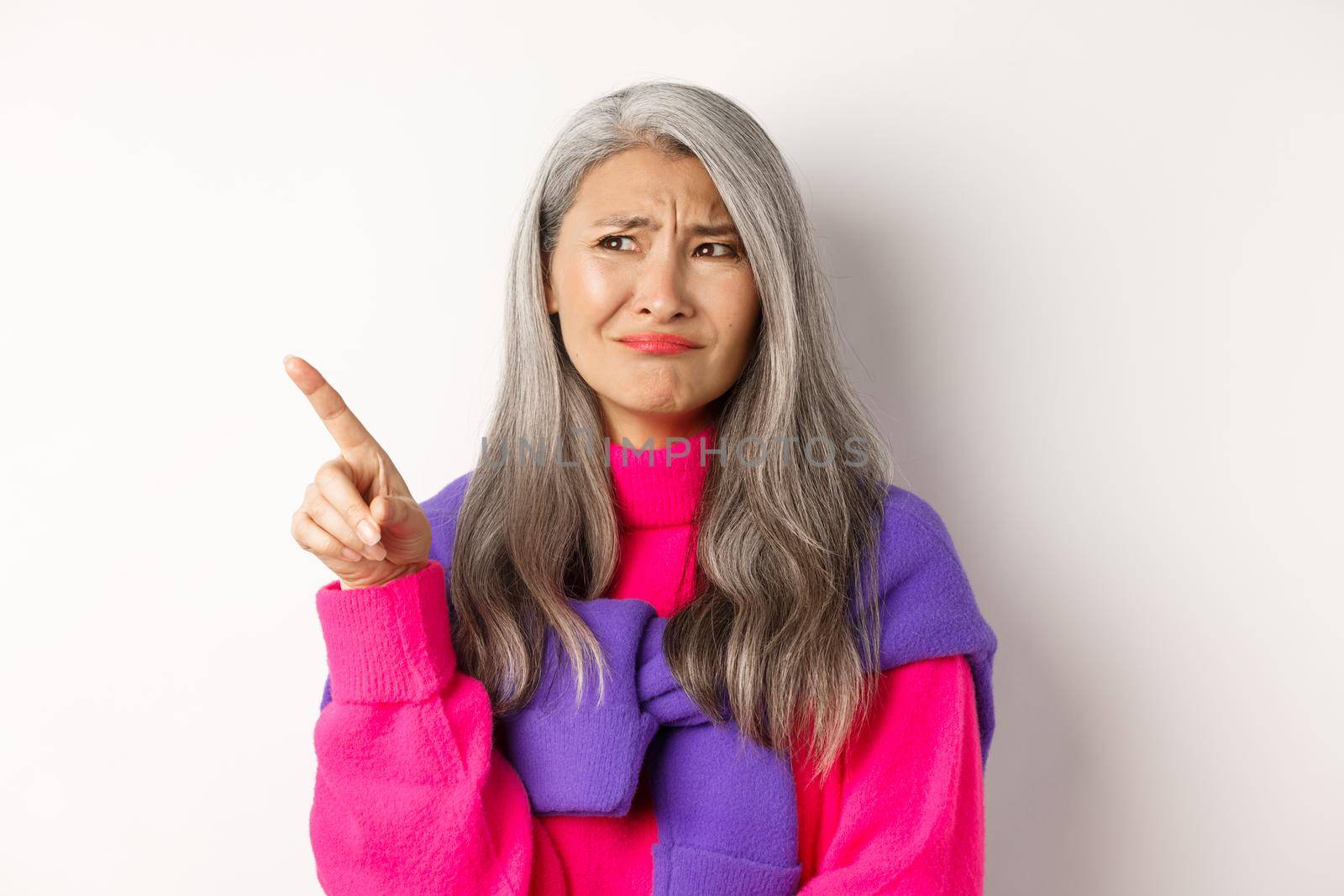 Displeased asian grandmother in pink sweater, grimacing and frowning skeptical, pointing finger upper left corner, standing over white background.