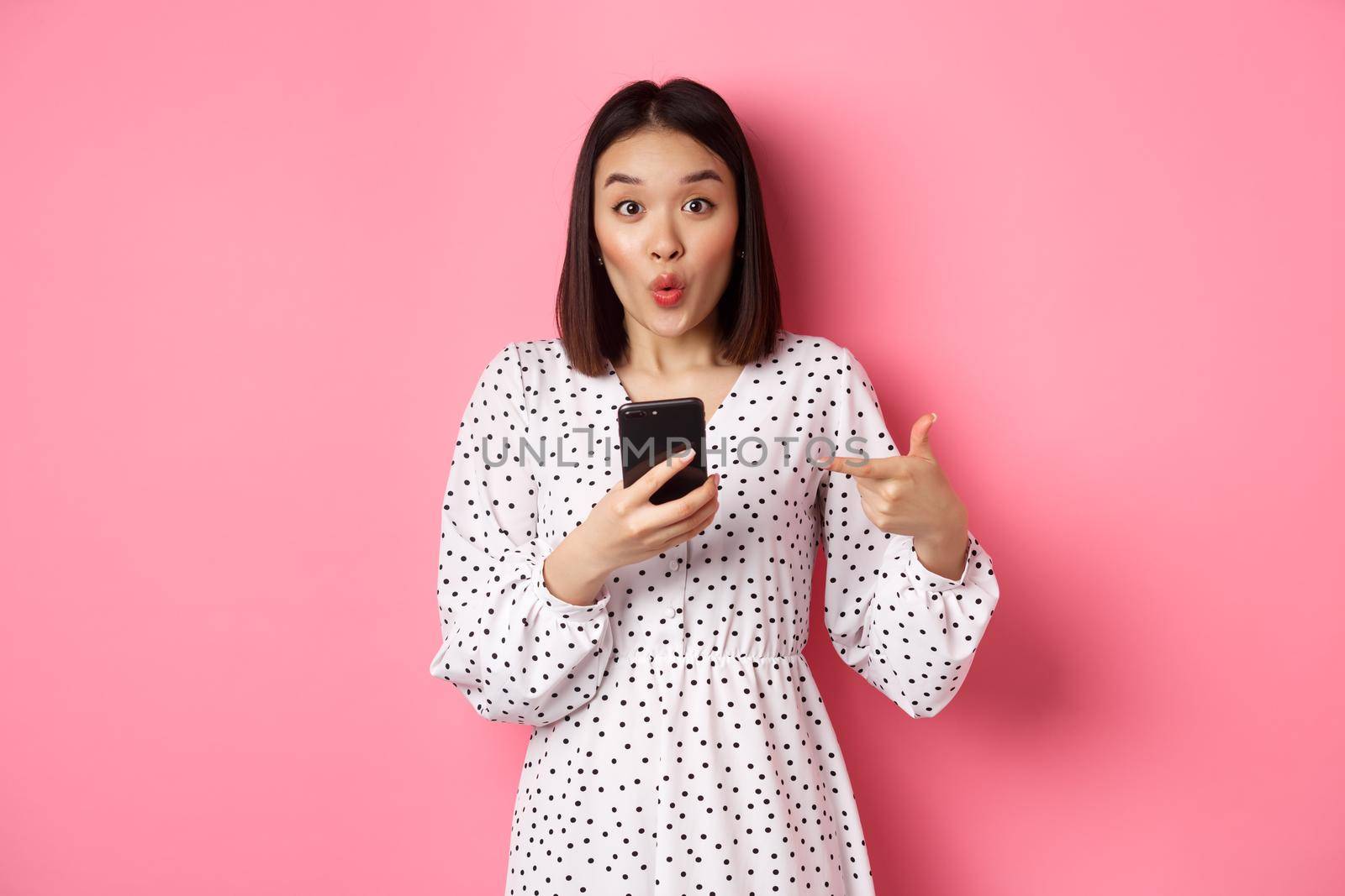 Online shopping and beauty concept. Amazed asian woman pointing finger at mobile phone, recommending smartphone app, standing over pink background.