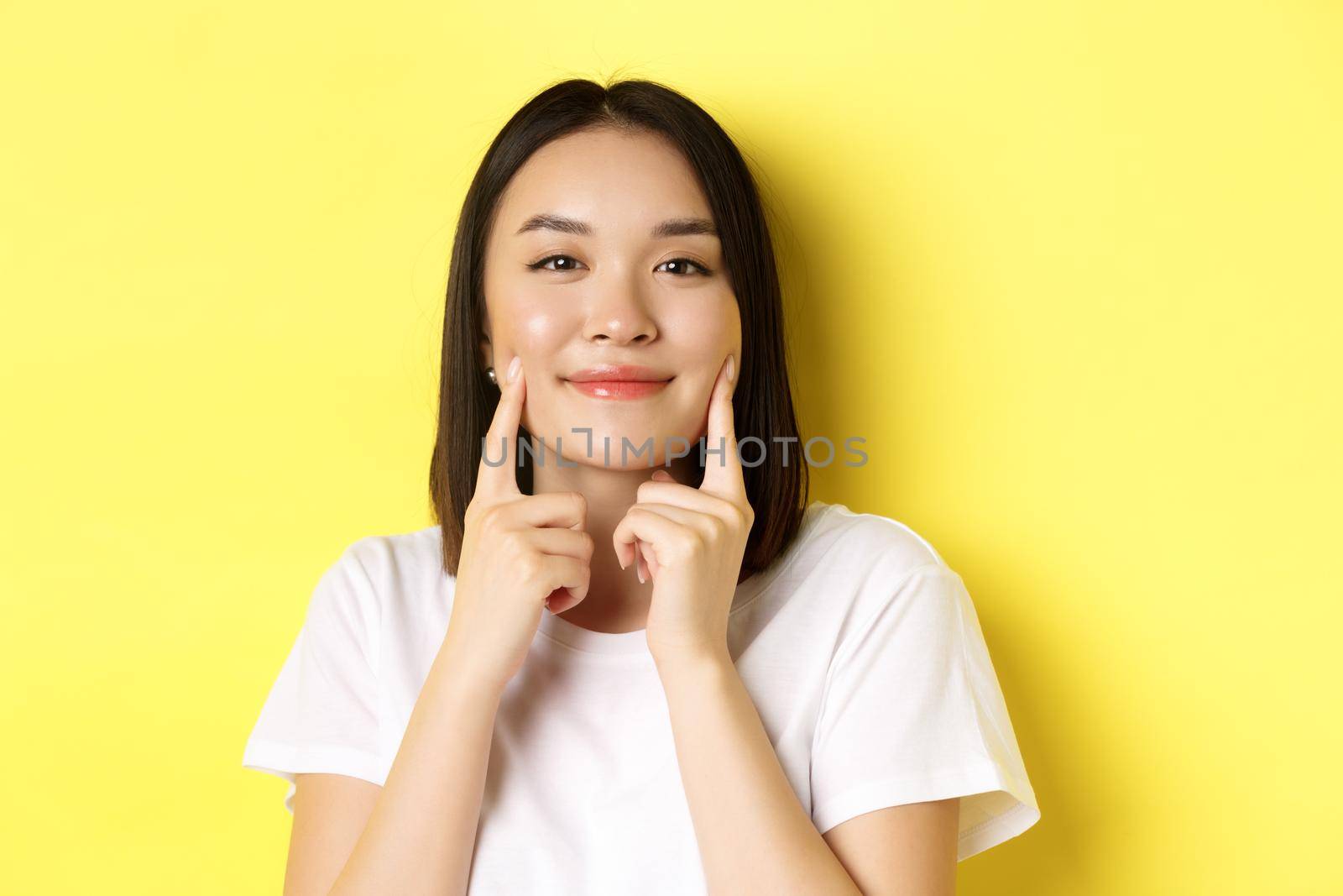 Beauty and skincare. Close up of young asian woman with short dark hair, healthy glowing skin, smiling and touching dimples on cheeks, standing over yellow background by Benzoix