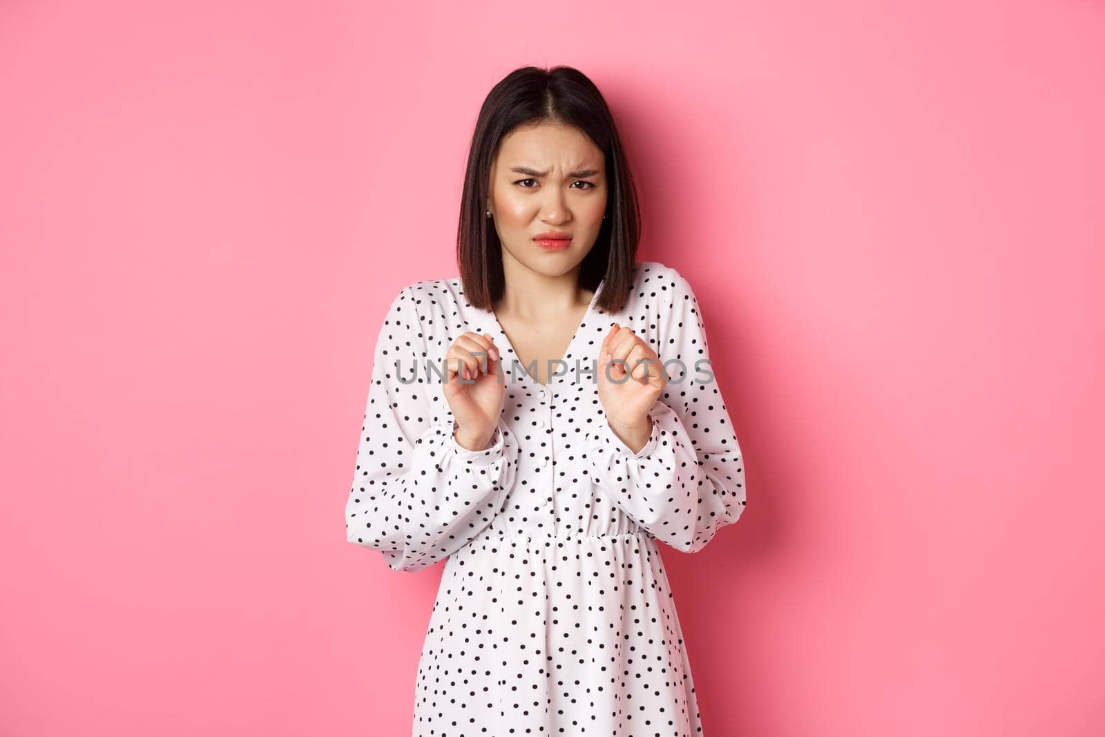 Disgusted asian woman staring with aversion and dislike, frowning and grimacing dissatisfied, standing in dress against pink background.