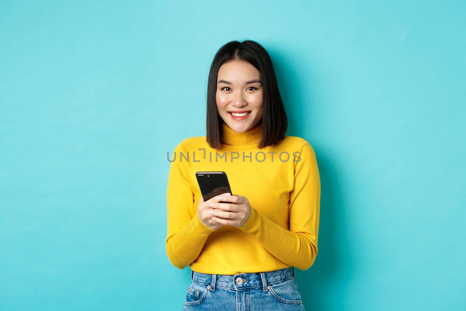 Beautiful and stylish asian woman shopping online on mobile phone, standing over blue background.