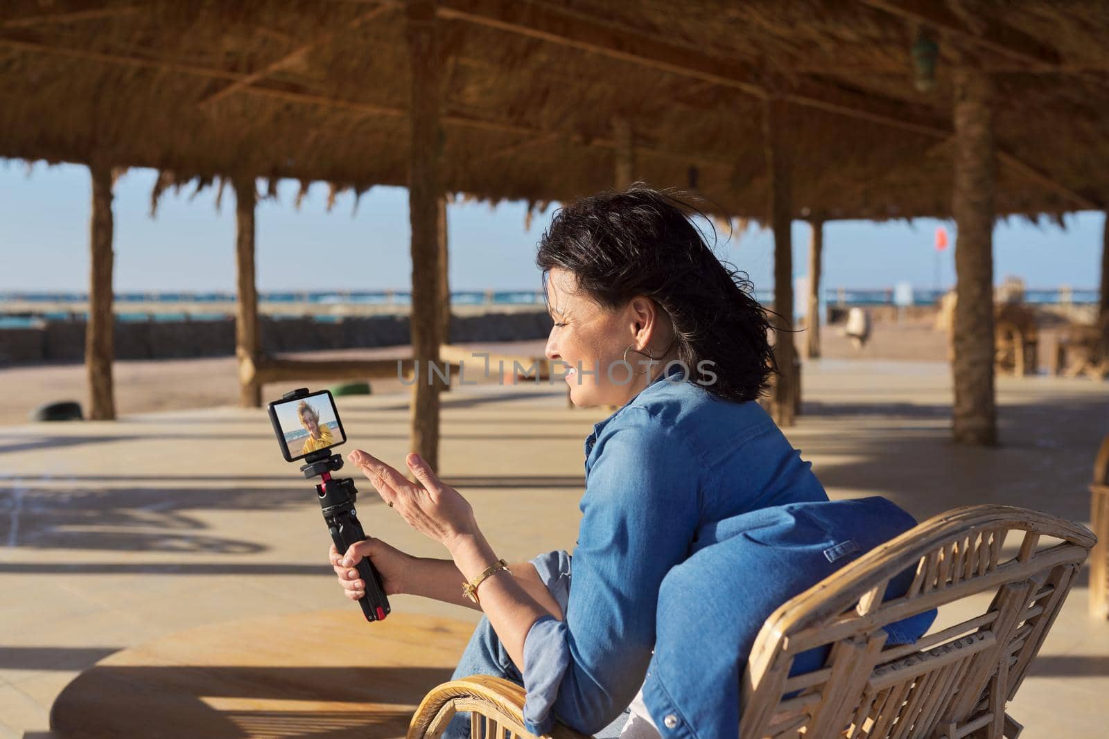 Woman at seaside resort in rattan chair talking to her female friend using video call on smartphone. Lifestyle, technology, telecommunications concept