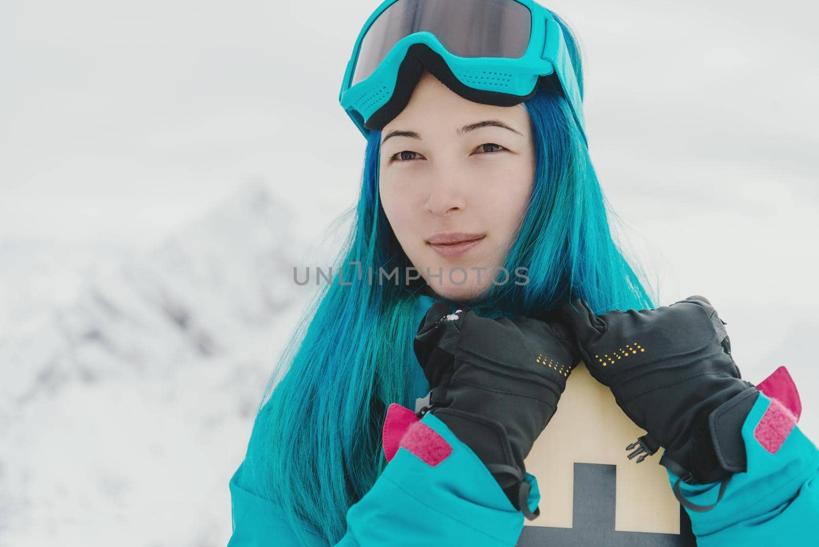 Smiling young woman with blue hair standing with snowboard on background of snowy mountains