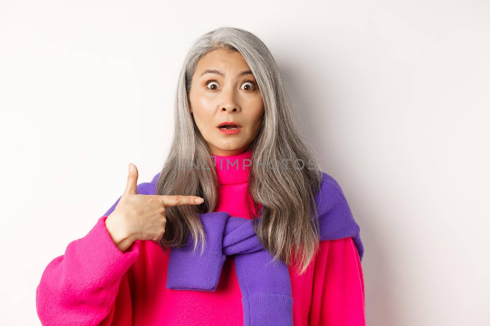 Confused asian senior woman with grey hair gasping startled, pointing at herself and looking at camera, standing over white background by Benzoix