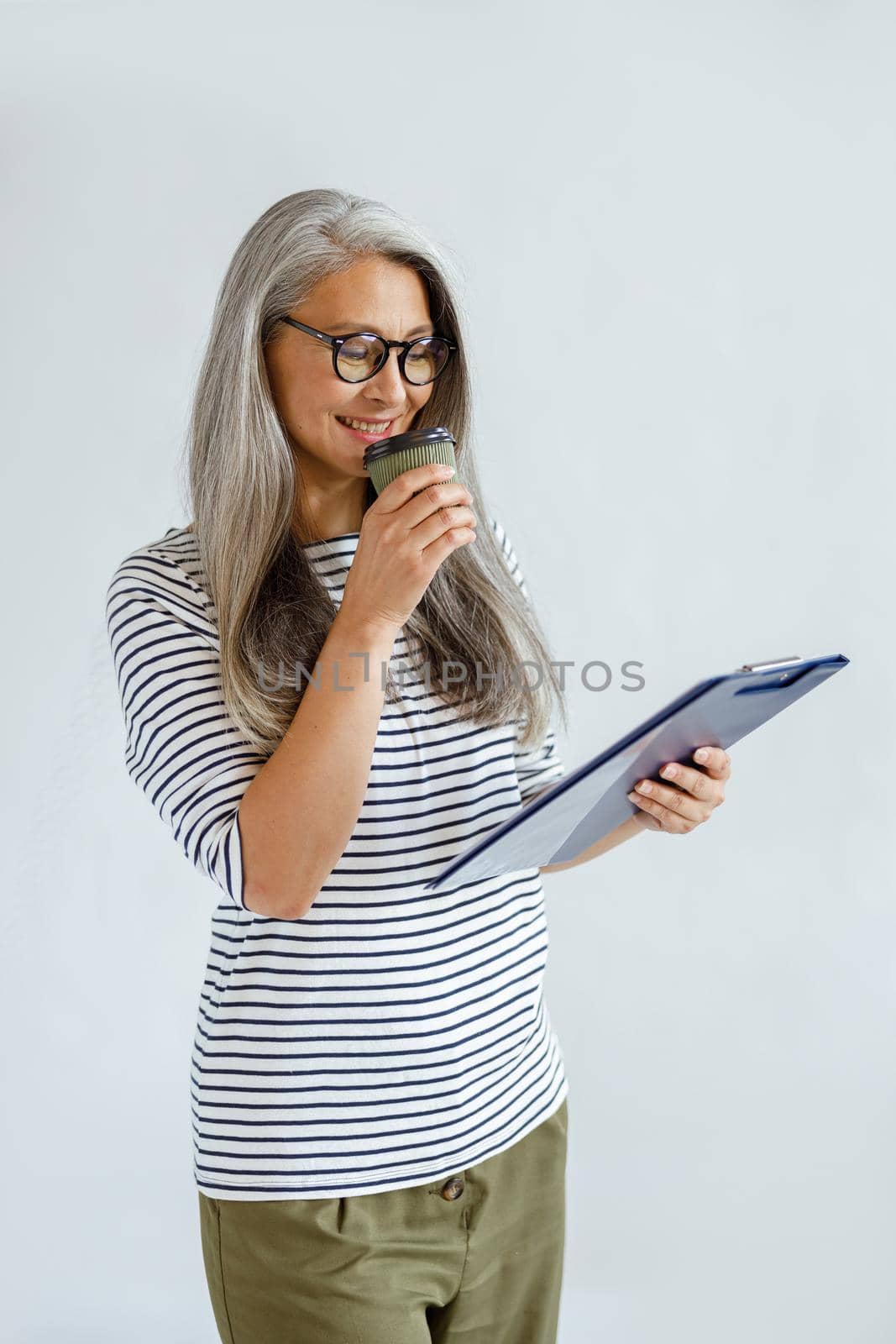 Smiling mature Asian lady with loose hair drinks coffee reading paper on clipboard standing on light grey background in studio