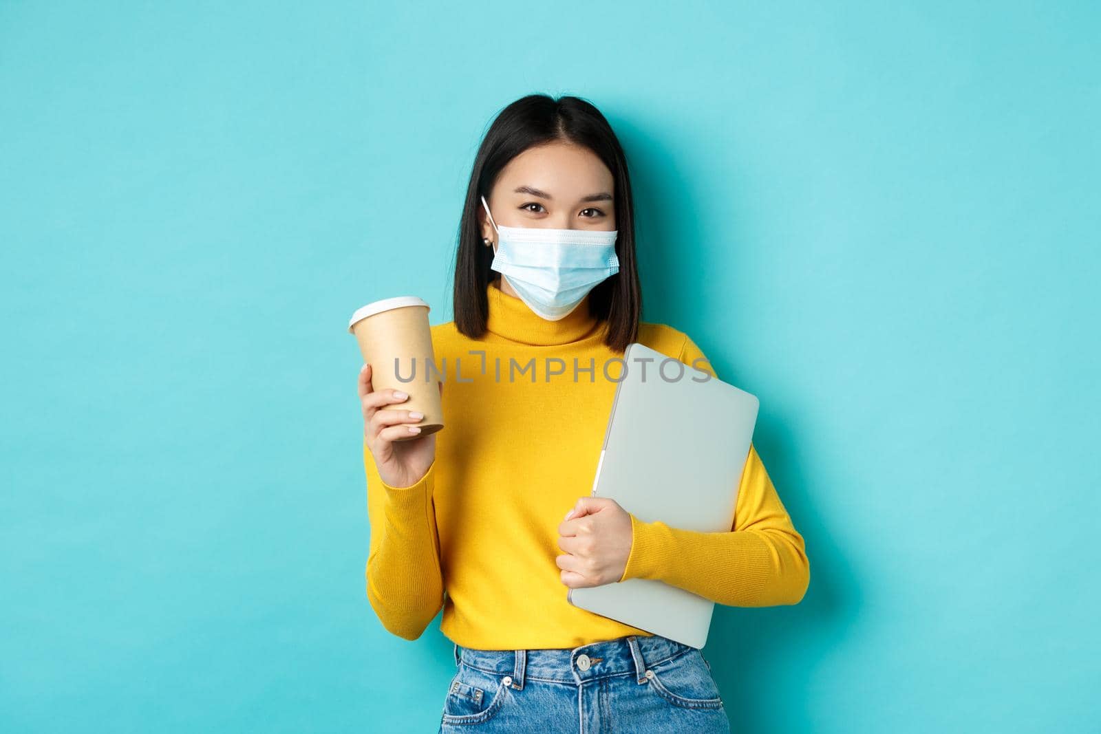 Covid, pandemic and social distancing concept. Stylish asian woman wearing medical mask, holding cup of coffee and laptop, going to work, standing over blue background by Benzoix