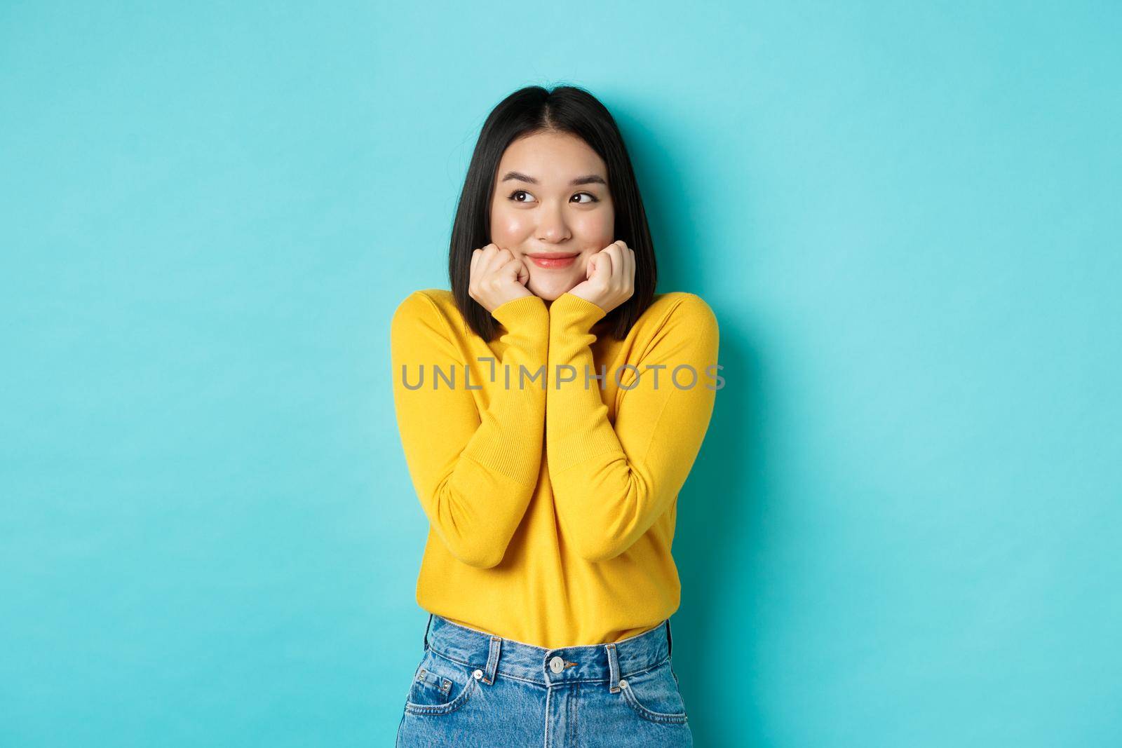 Beauty and fashion concept. Beautiful asian woman blushing and smiling, looking dreamy left, imaging something cute, standing against blue background.