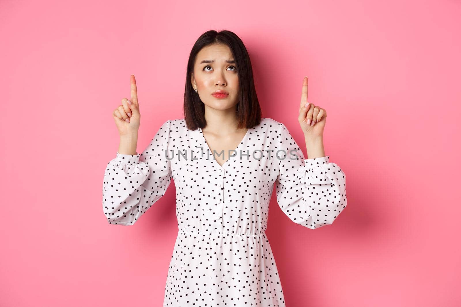 Worried and concerned asian girl in cute dress frowning, pointing and looking up with sad face, standing over pink background by Benzoix