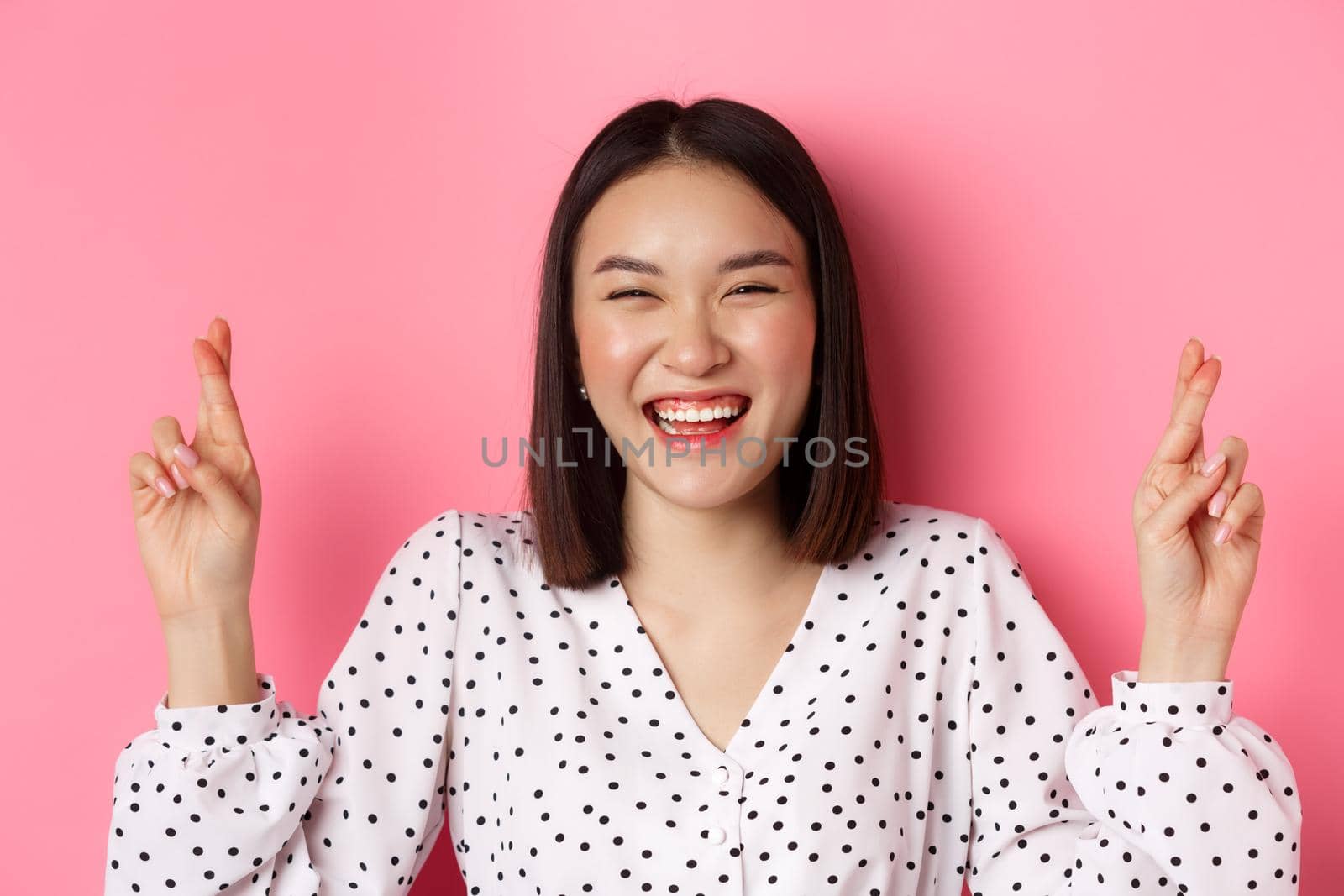 Beauty and lifestyle concept. Close-up of happy asian woman making a wish, holding fingers crossed for good luck and smiling optimistic, standing over pink background.