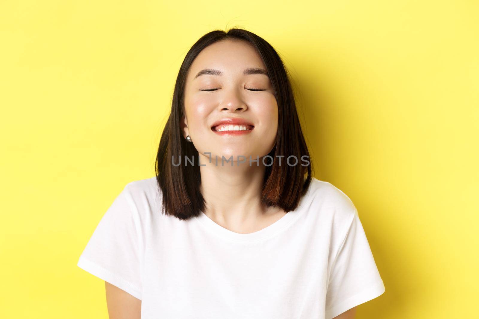 Close up of happy and relaxed asian woman enjoying sun, smiling with eyes closed and looking joyful, standing over yellow background by Benzoix