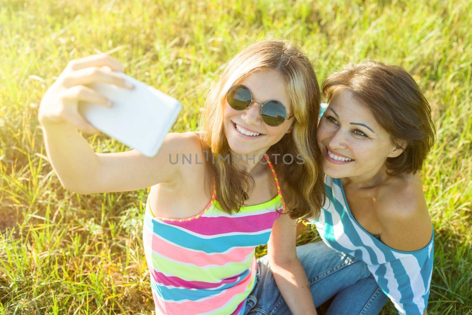 Portrait of beautiful adult mother and her daughter teenage girl making a selfie using a smart phone and smiling. Photo taken in nature, summer day