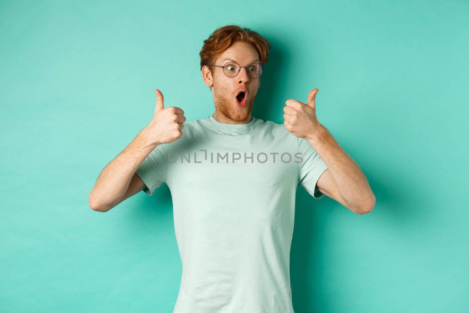 Excited young man with red hair and beard, staring at promo with awe, showing thumbs-up in approval, praising offer, standing over turquoise background.
