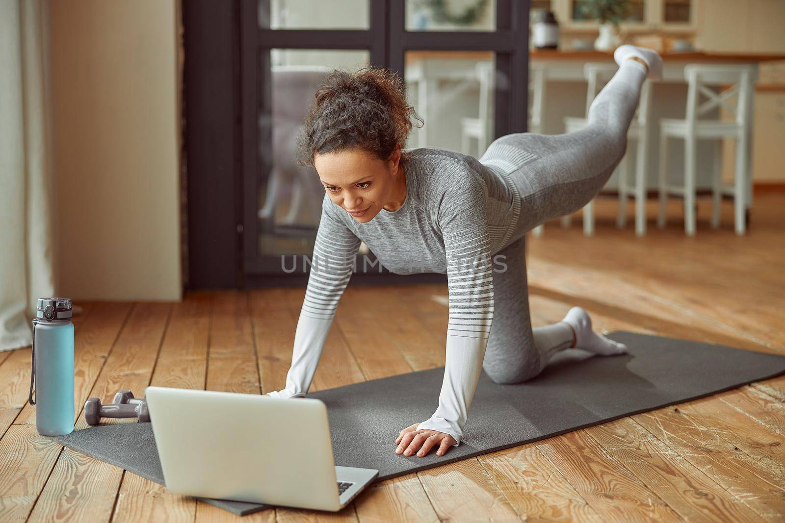 Cheerful slim woman is doing exercises on mat while watching training on laptop at home